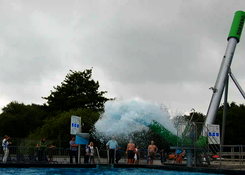 Personen schauen auf einen grünen Cannonball vor einem Schwimmbecken, aus dem Wasser sptitzt