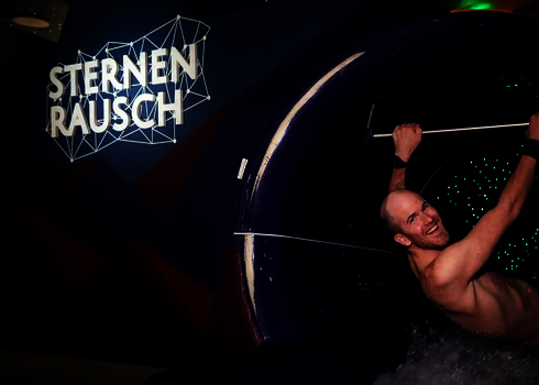 Ein lachender Mann sitzt im Startbereich einer dunklen Wasserrutsche mit LED-Lichteffekten.