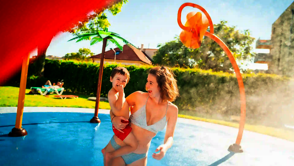 Eine Frau mit Kind auf dem Arm auf einem Wasserspielplatz mit Wie-Spray Elementen.