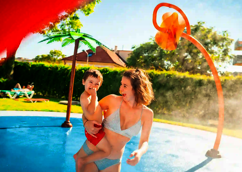 Eine Frau mit Kind auf dem Arm auf einem Wasserspielplatz mit Wie-Spray Elementen.