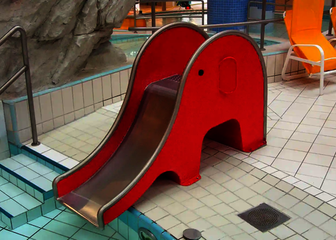  Rote Rutsche in Elefantenform steht am Rand eines flachen Wasserbeckens im Kinderbereich eines Schwimmbades.