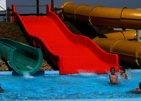 Eine rote GFK Breitwellenrutsche neben einer gelben und einer blauen Wasserrutsche in einem Freibad mit Badegästen.