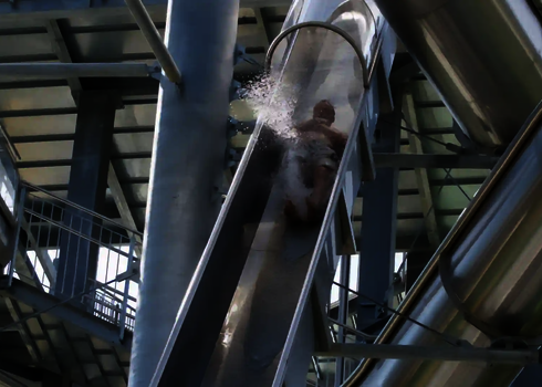 Person rutscht in liegender Haltung in einer steilen, transparenten Freifallrutsche aus Edelstahl.