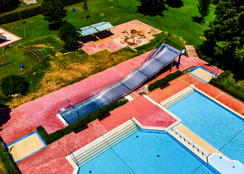Vogelperspektive auf ein Freibad mit Blick auf eine Breitwellenrutsche und mehrere Schwimmbecken.