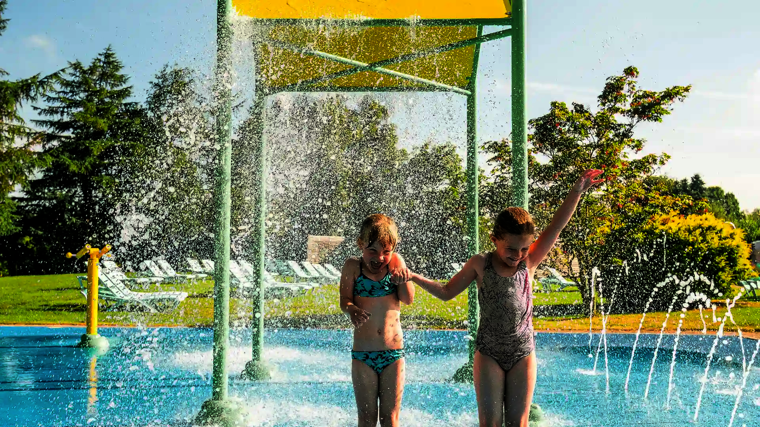 Zwei Kinder halten sich lachend an den Händen und stehen im spritzendem Wasser einer Wasserspielstruktur.