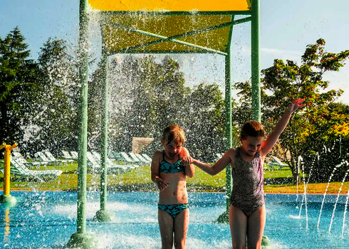 Zwei Kinder halten sich lachend  an den Händen und stehen im spritzendem Wasser einer Wasserspielstruktur.