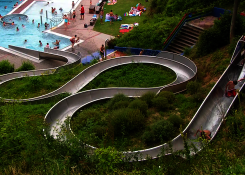 WR1040-Rutsche im Außenbereich mit S-Kurvenführung durch begrünte Böschung, Freibad im Hintergrund.
