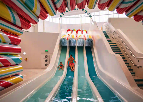 Multilane im Hallenbad, ein Mann und ein Kind rutschen nebeneinander ins Wasser.