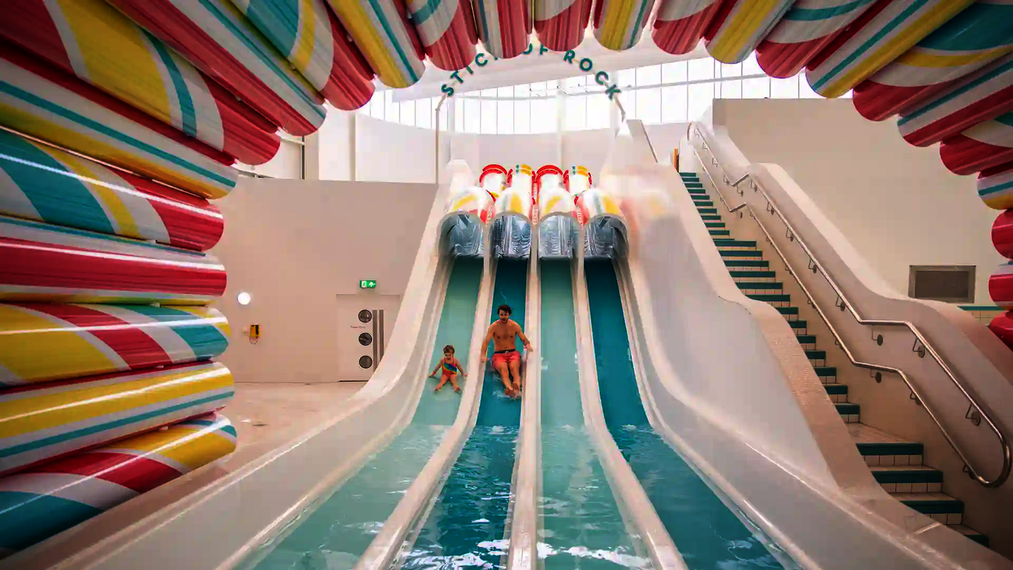 Multilane im Hallenbad, ein Mann und ein Kind rutschen nebeneinander ins Wasser.