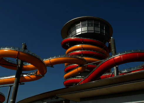 Spiralförmige gelbe und rote Röhre der WR1040-Rutsche vor blauem Himmel und Glasfassade.