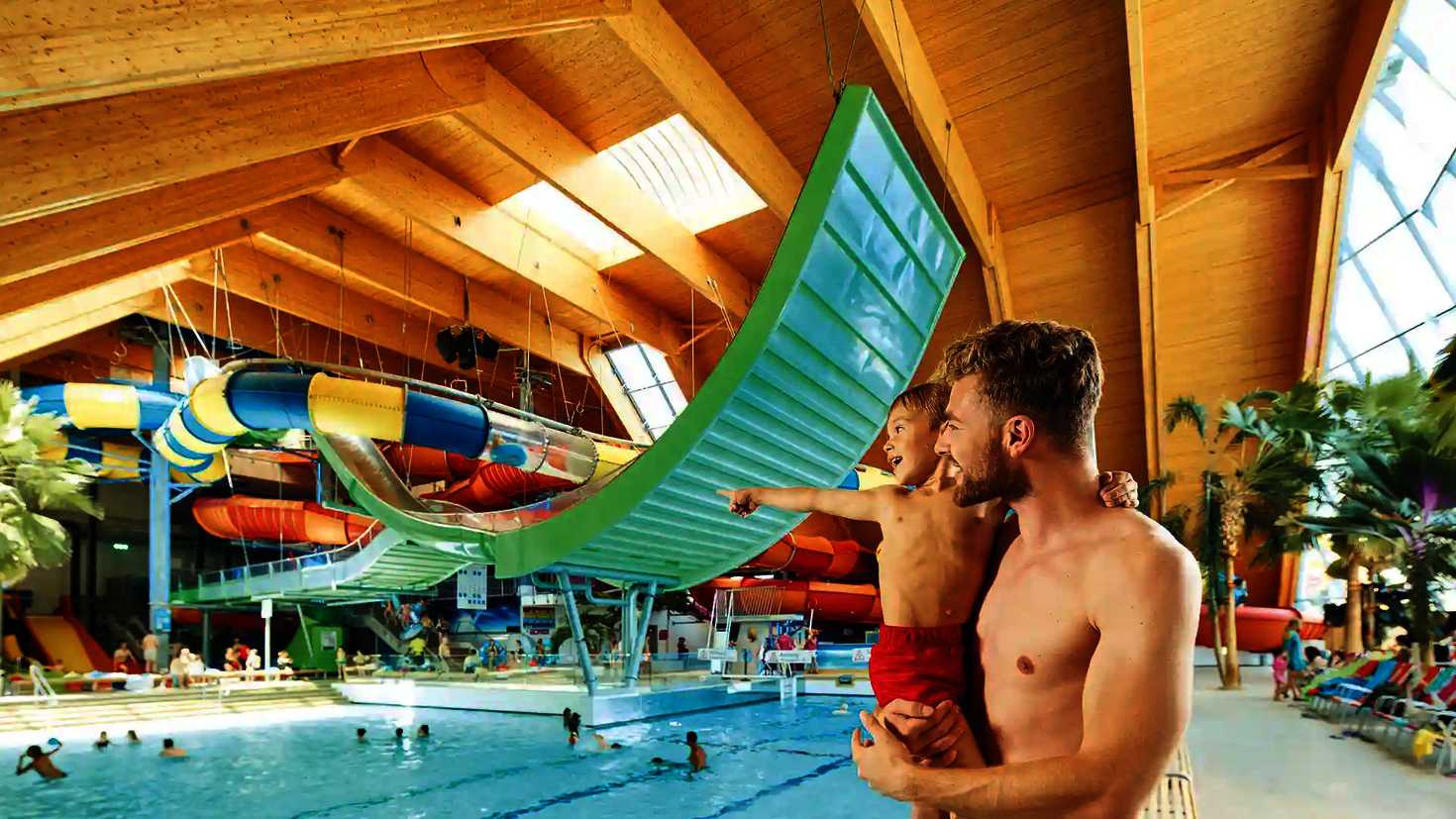Vater mit Kind im Arm zeigt auf die große grüne Boomerang-Rutsche über dem Wasserbecken.