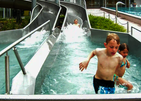 Mehrere Kinder rutschen aus einer Rutsche ins Wasser, eines läuft lachend weg.