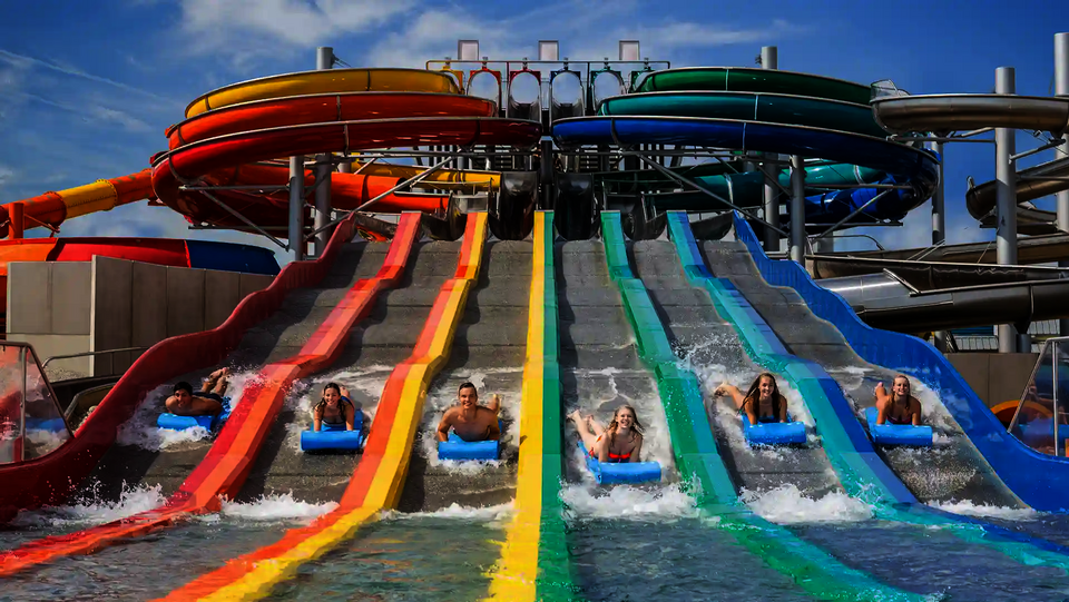 Sechs Personen auf blauen Rutschmatten rutsche eine bunte Matracer Wasserrutsche herunter.