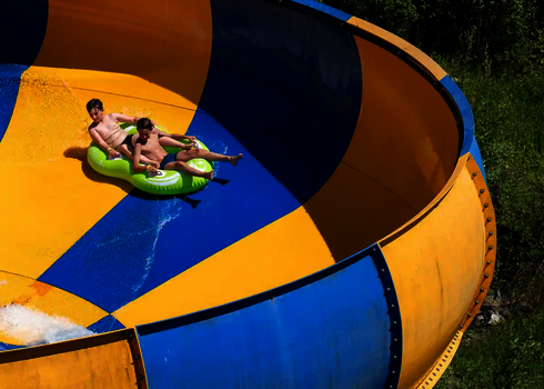 Zwei Kinder rutschen hintereinander sitzend in einem grünen Reifen auf einer Wasserrutsche.