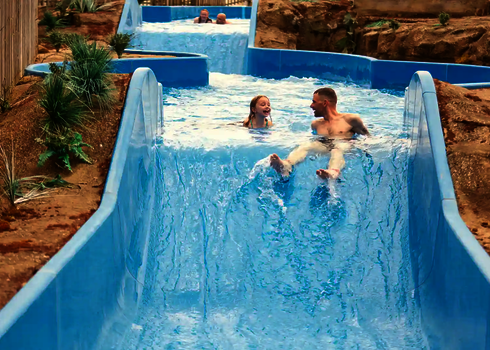 Mehrere Personen rutschen lachend den Wildwasserfluss entlang, sandige Umgebung.