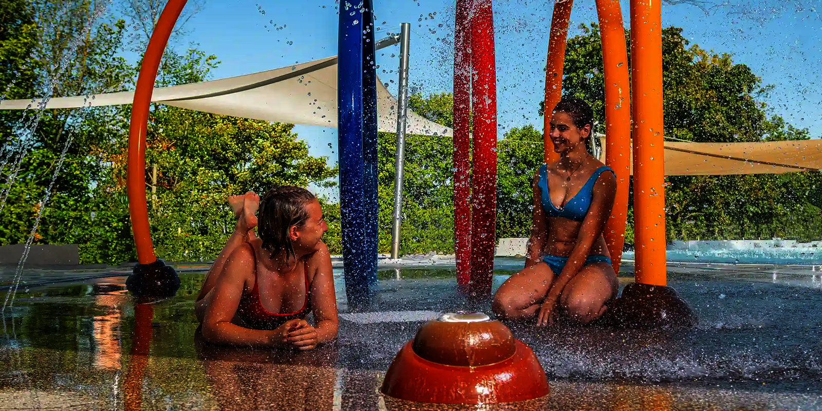 Zwei Frauen entspannen im Wasserspielbereich unter bunten Wasserspritzbögen