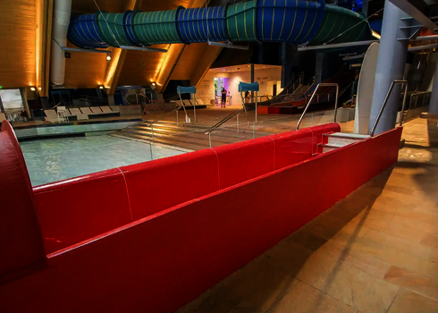 Innenansicht eines Hallenbads mit einem roten Auslaufbecken im Vordergrund und einer blau-grünen Röhrenrutsche im Hintergrund.