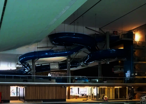 Blauer Crazy River in überdachter Schwimmhalle, getragen von Stützen und Stahlseilen.