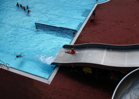 Ansicht von oben auf einen Teilbereich eines Schwimmbades, mit einer Breitwellenrutsche auf der eine Person ruscht.