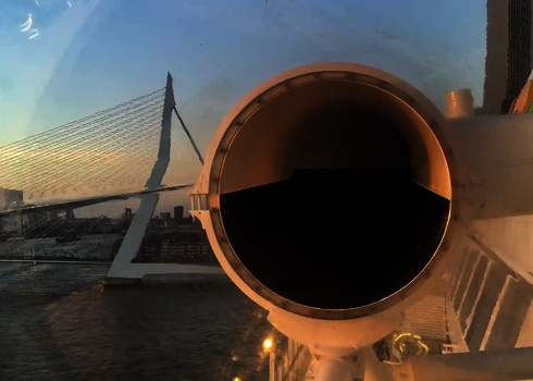 Blick aus einer transparenten Röhre einer Wasserrutsche mit Stadtpanorama und Brücke bei Sonnenuntergang.