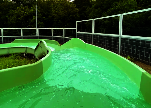 Grüne Rutschbahn mit Wasserströmung und Kurve, umgeben von einem Sicherheitsgeländer.