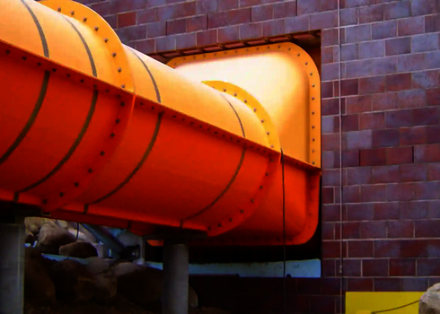 Orangefarbene Wasserrutsche durchdringt rechteckige Öffnung in dunkler Ziegelfassade.