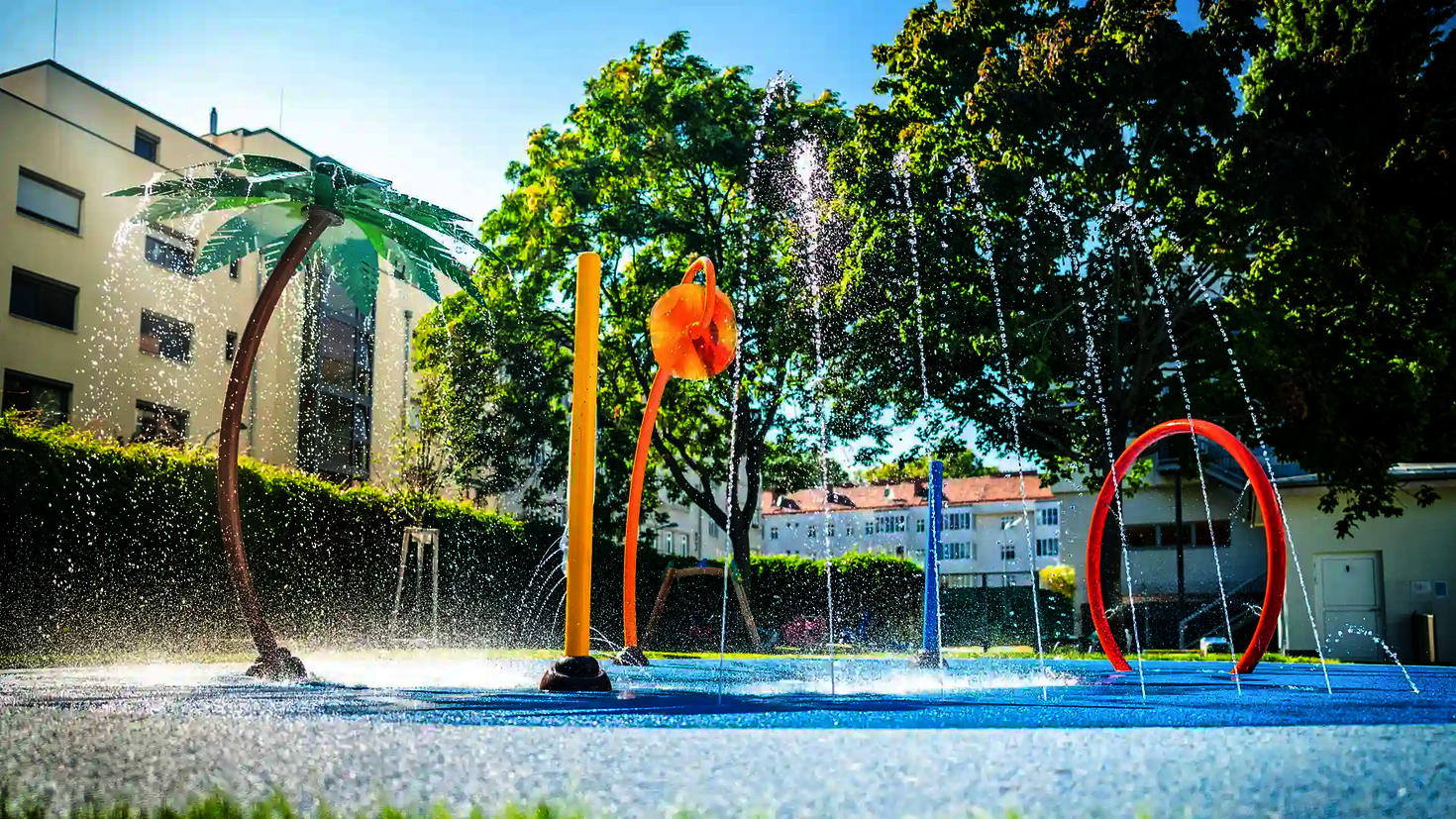 Wasserspielplatz mit spritzenden Wie-Spray Elementen vor Hotelanlage.
