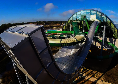 Blick auf die Hinterseite einer Boomerang Wasserrutsche, im Hintergrund befindet sich ein grün weißer Family Twister und ein Fallstart vor einem Hallenbad.
