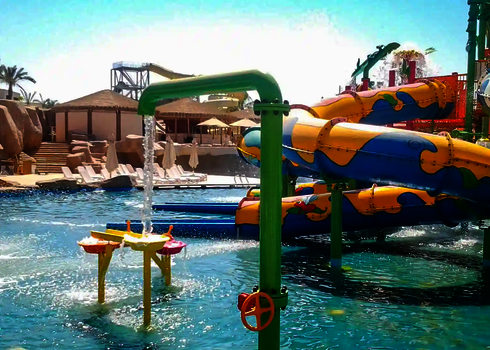 Wie-Spray Elemente und blau-gelbe RR900-Kids Rutsche in einem Wasserpark mit Palmen im Hintergrund.