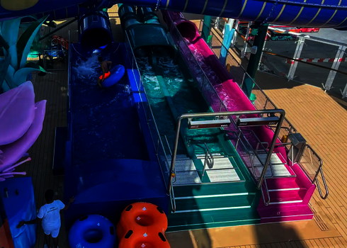 Kind mit blauem Rutschreifen in einem Auslauf neben der bunten 2-in-1 Racer Wasserrutsche. 