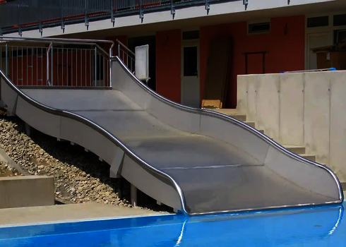 Eine kurze Breitwellenrutsche vor einem Gebäude, die in ein Schwimmbecken endet.