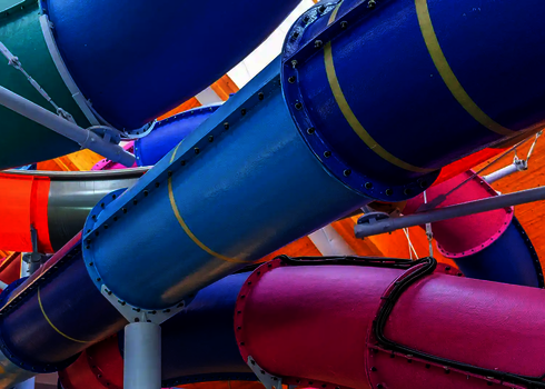 Blick nach oben auf verschlungene, bunte Wasserrutschen in einer überdachten Anlage.
