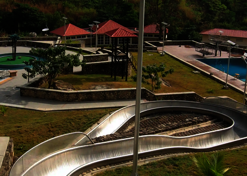 Edelstahlrutsche mit enger Kurve verläuft durch Parkanlage neben einem Freibad.