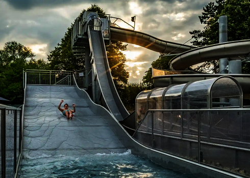 Eine Person rutscht bei Sonnenuntergang auf einer Breitwellenrutsche, daneben befindet sich ein Freifall. 