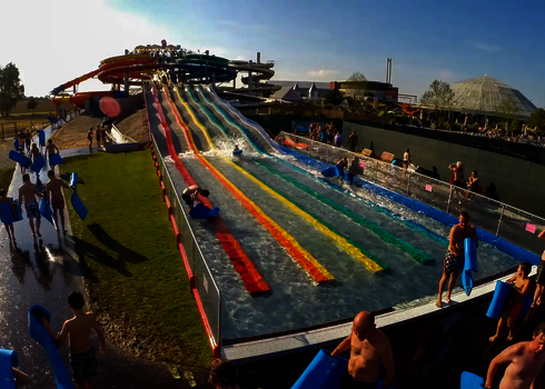 Besucher gehen mit blauen Matten zur Wasserrutsche Matracer, bunte Röhren im Hintergrund.