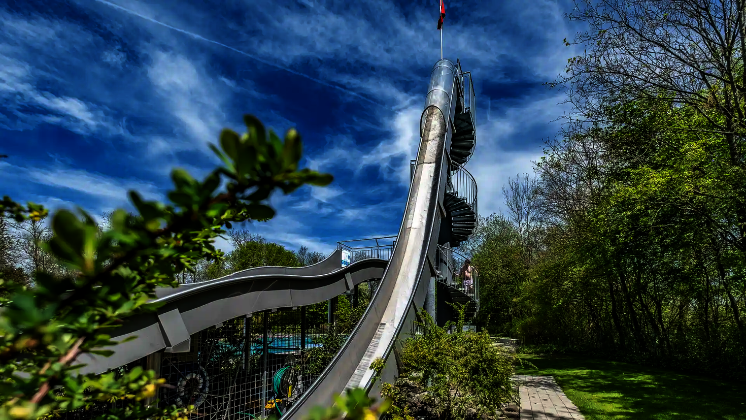 Steile Freifallrutsche mit Wendeltreppe im Freibad, im Hintergrund ein blauer Himmel.