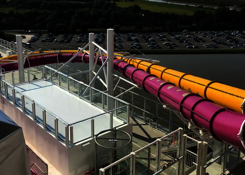 Zwei Wasserrutschen in Gelb und Pink verlaufen auf einem Kreuzfahrtschiff vor einem Hafen.