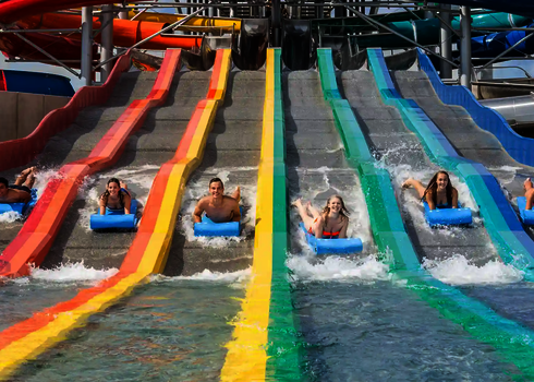 Six people slide down a matracer water slide in the water park on mats next to each other.