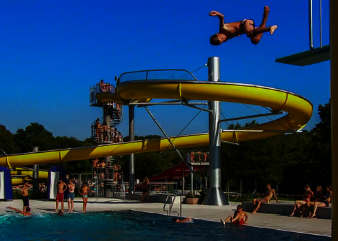 Junge springt vom Sprungturm ins Becken, Gäste beobachten vom Rand.