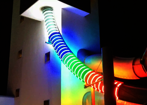 Außenansicht einer beleuchteten Röhrenrutsche mit farbigen LED-Ringen bei Nacht.