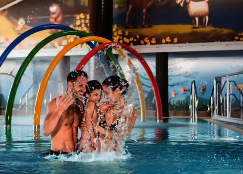Familie steht lachend in einem Wasserbecken, dahinter das Wie-Spray Bauteil "Regenbogen".