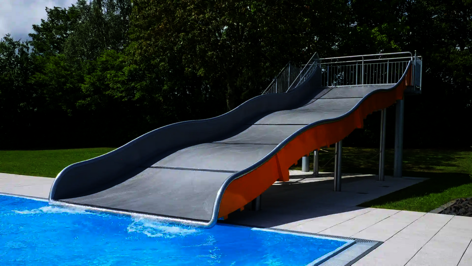 Eine Edelstahl Breitwellenrutsche mit orangefarbenen Seitenwänden vor einem Schwimmbecken stehend, im Hintergrund grüne Bäume.