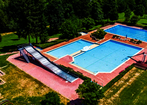 Vogelperspektive auf ein Freibad umgeben von Bäumen, mit mehreren Schwimmbecken und einer Breitwellenrutsche.