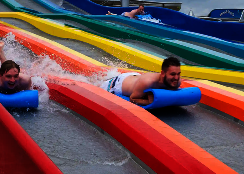 Two men slide side by side on blue mats down a coloured matracer water slide in a water park.