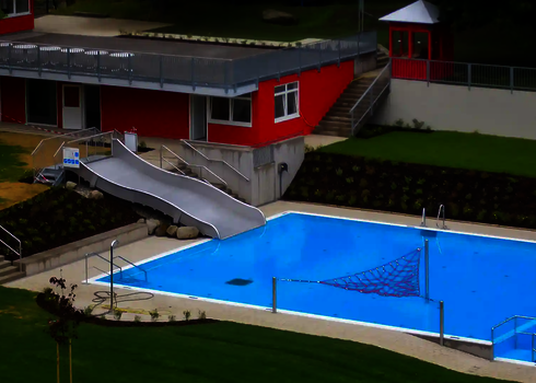 Blick auf ein Freibad mit einer Breitwellenrutsche, einem blauen Schwimmbecken und einem roten Gebäude im Hintergrund.