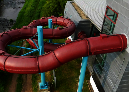 Luftaufnahme einer roten, gedämmten Röhrenrutsche mit engen Kurven neben einem Bachlauf.