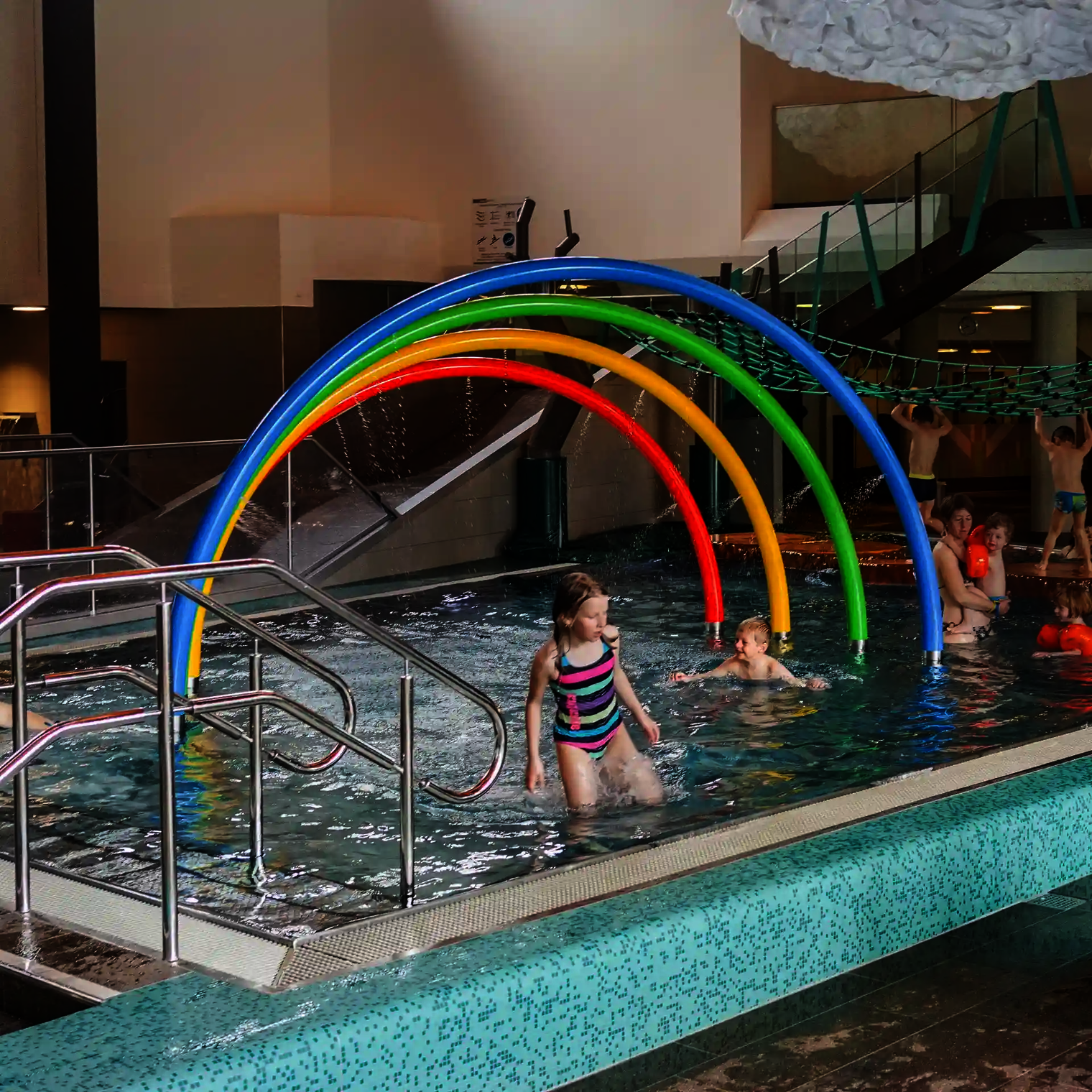 Kinder in Wasserbecken spielen unter dem Wie-Spray Element "Regenbogen".