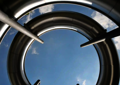 Blick von unten auf eine kreisförmige Edelstahlrutsche vor blauem Himmel.