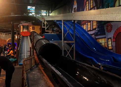 Indoor-Spielplatz mit zwei blauen und einer schwarzen Rutsche, am rechten Rand befinden sich Personen.