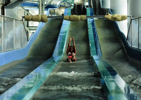 Frau rutscht allein eine breite offene Wasserrutsche in einem hellen Innenbereich hinab.